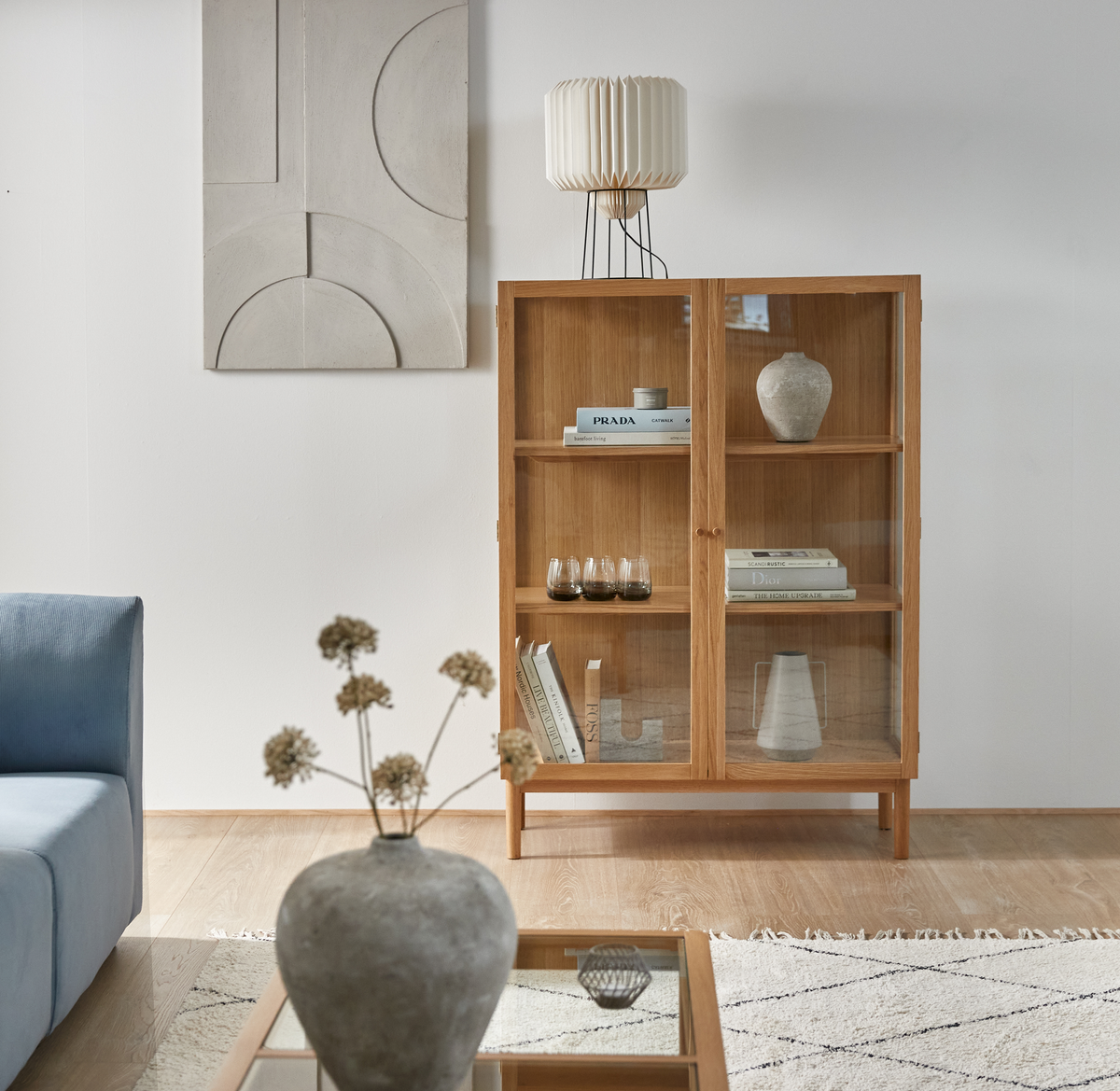 Ljus skandinavisk vardagsrumsstyling med vitrinskåp i trä, boktitlar PRADA och DIOR på hyllorna, blå soffa, vas på soffbord och veckad bordslampa.