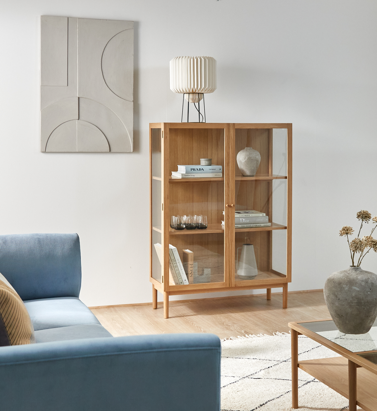Ljust vardagsrum med vitrinskåp i ek med glasdörrar; hyllor med böcker som Prada och Dior, glas och keramik; blå soffa, glasbord och ljus matta.