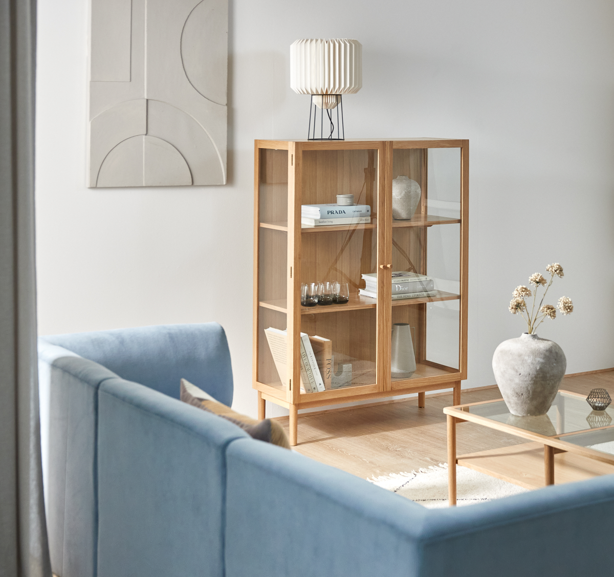 Ljus vardagsrumsinteriör med ljusblå soffa, glas- och träsoffbord och vitrinskåp i ek med böckerna PRADA och Dior samt en veckad bordslampa; minimalistisk dekoration och väggkonst.