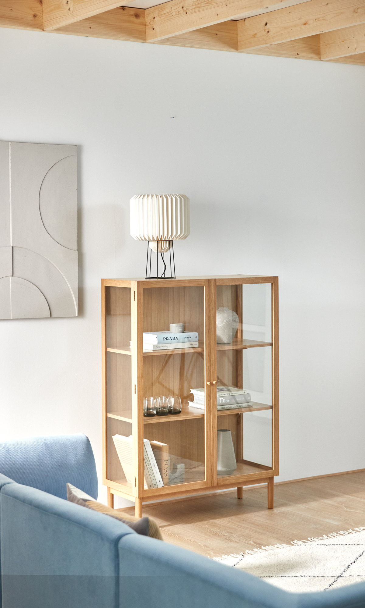 Ljust skandinaviskt vardagsrum med vitrinskåp i ek med glasdörrar, stylat med böcker som Prada och Dior, små glas och keramik, med vit veckad bordslampa ovanpå, blå soffa och ljus matta.