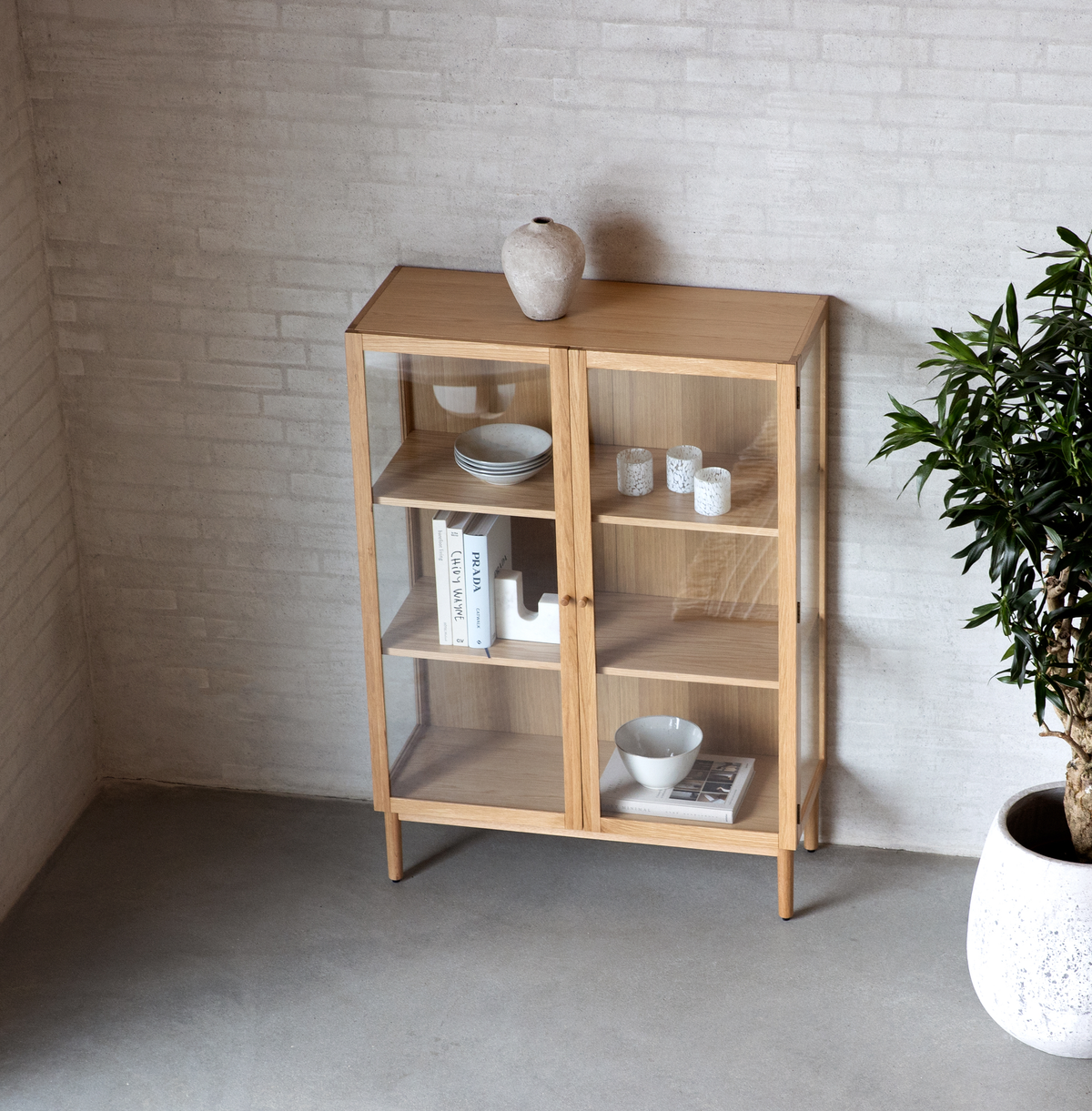 Vitrinskåp i ljus ek med glasdörrar, stylat med tallrikar, skålar, små ljus och böcker – varav en med titeln PRADA – i en minimalistisk interiör med krukväxt.