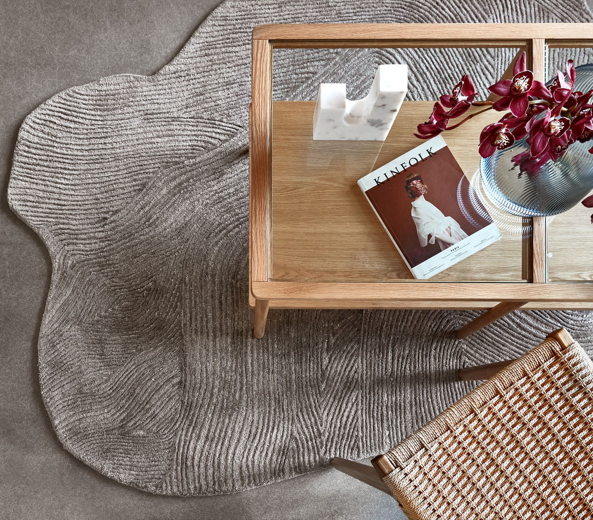 Top-down av ett soffbord i trä och glas på en texturerad grå matta, med en Kinfolk-tidning, en vit marmordekoration, en ribbad glasvas med mörkröda orkidéer och en stol med flätad sits.