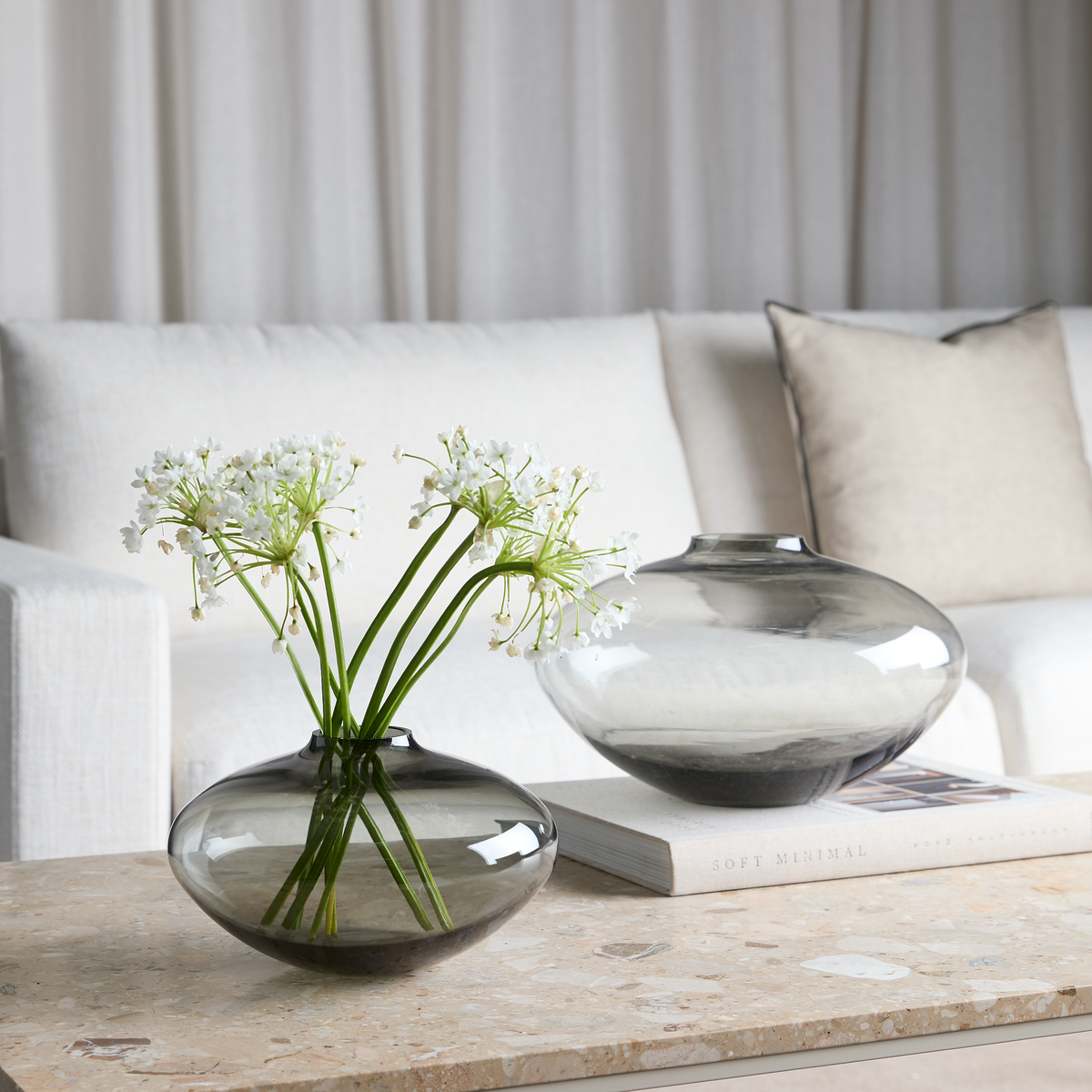 Två rökgrå glasvaser på ett stenbord framför en ljus soffa; den främre med vita blommor, den bakre står på boken “Soft Minimal”.
