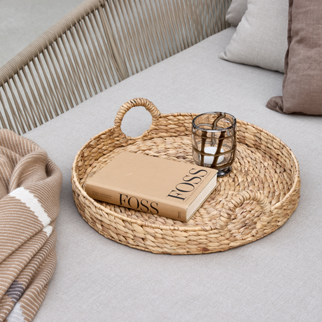 Flätad rund bricka på ljusgrå soffa med en bok med titeln FOSS och en glaslykta med ljus, intill en beige randig pläd och kuddar.