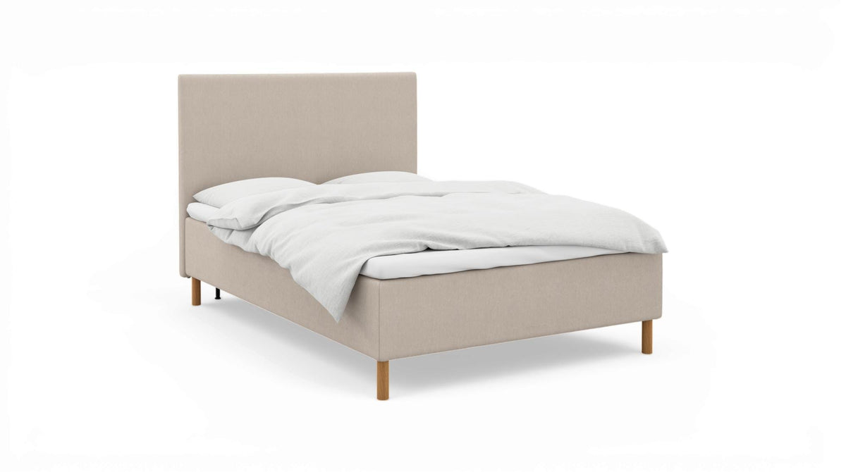 Beige stoppad säng med vit bäddning och träben, frilagd.