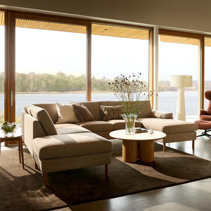 Ljust vardagsrum med beige modulsoffa, runt soffbord i trä och stora panoramafönster med sjöutsikt.