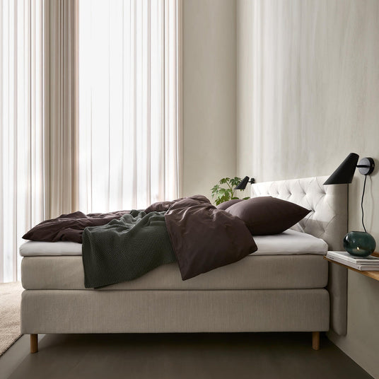 Ljust sovrum med beige kontinentalsäng och vadderad sänggavel, mörkbruna sängkläder och grön pläd, svarta vägglampor, skira gardiner samt liten hylla med glasvas och böcker.