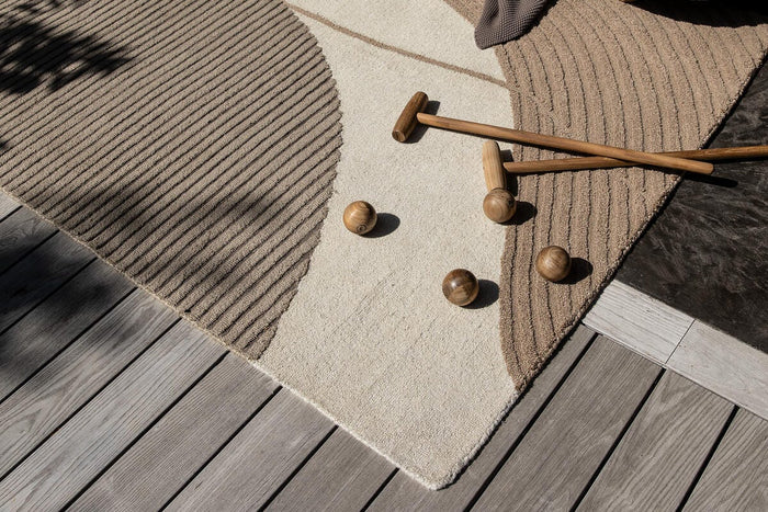 Beige och taupefärgad abstrakt matta på en träterrass med böjd soffa, pläd samt träklubbor och kulor i solsken.