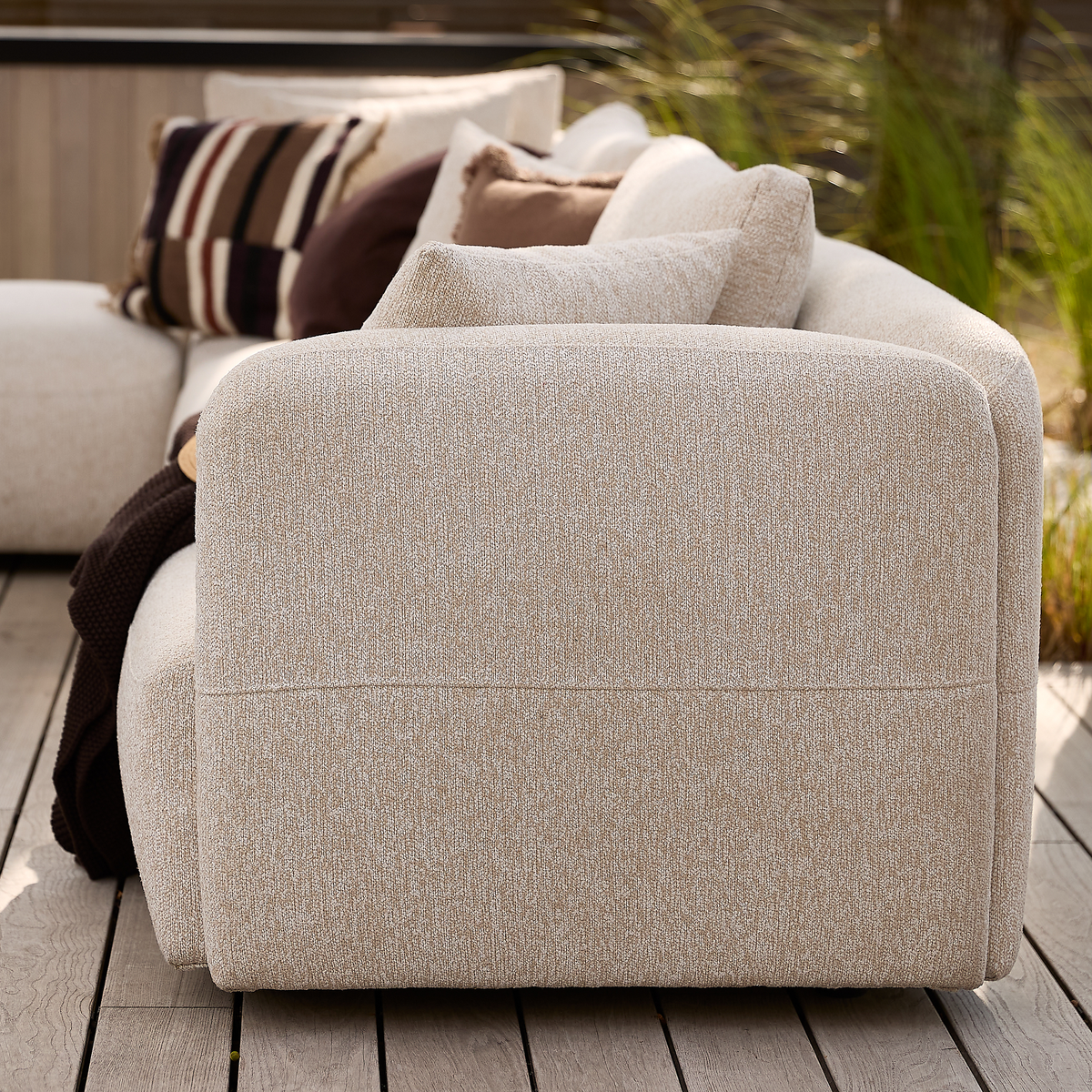 Beige modulsoffa på trädäck med neutrala kuddar och brun pläd, utomhus i dagsljus.