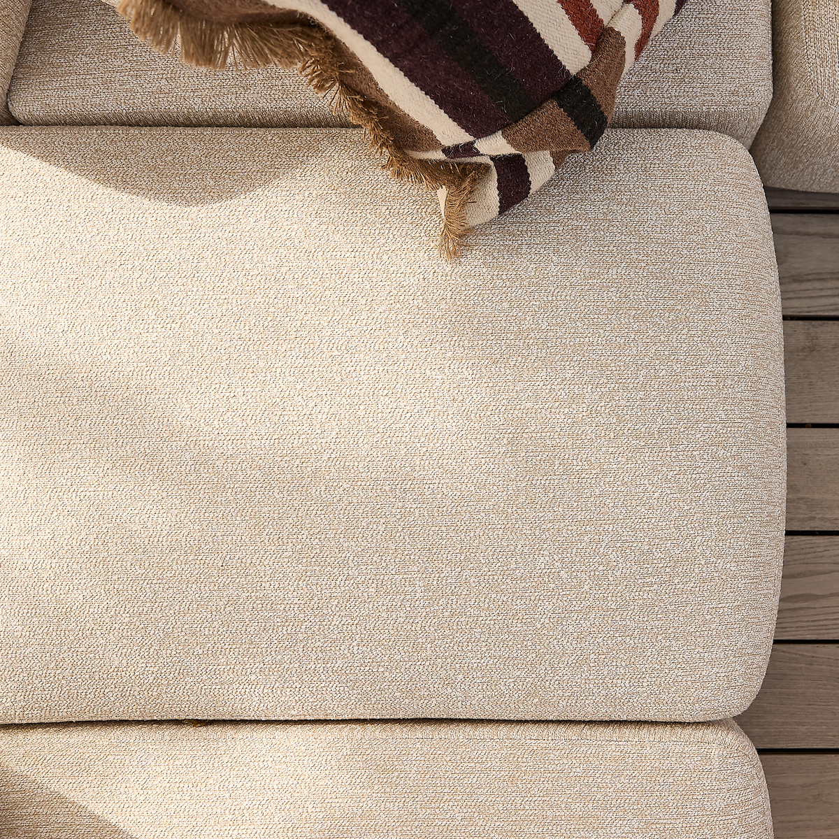 Närbild av beige, strukturerad soffdyna med en randig pläd på ett trädäck – utomhus sittplats i dagsljus.