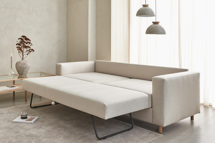Beige bäddsoffa utdragen i minimalistiskt vardagsrum med två pendellampor, skira gardiner och glasbord med vas och ljus.
