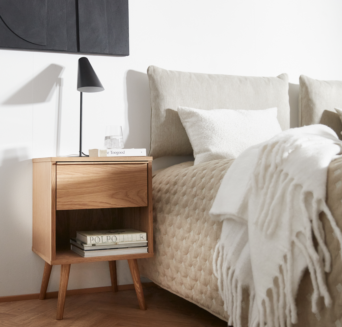 Sovrum med nattygsbord i trä, svart bordslampa, glas vatten och böckerna ”Faye Toogood” och ”POLPO”; beige bäddning med vit pläd med fransar.