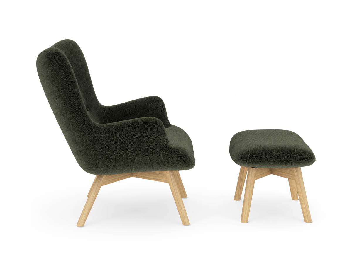 Mörkgrön fåtölj med matchande fotpall i mjuk textil och ben i ljust trä, sedd från sidan.