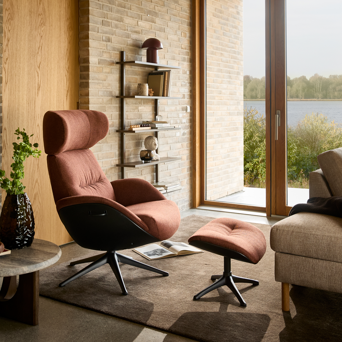 Brunröd recliner‑fåtölj med matchande fotpall i ett modernt vardagsrum, stora fönster mot en sjö, hyllsystem med lampa och böcker, beige soffa och brun matta.