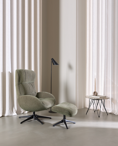 Loungefåtölj med fotpall i grön textil, svart golvlampa och litet sidobord med ljus och bok i ett ljust rum med gardiner.