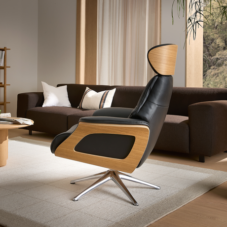 Modern loungestol i svart läder med trädetaljer och metallfot i ett ljust vardagsrum med brun soffa, kuddar, gardiner, matta och växter.