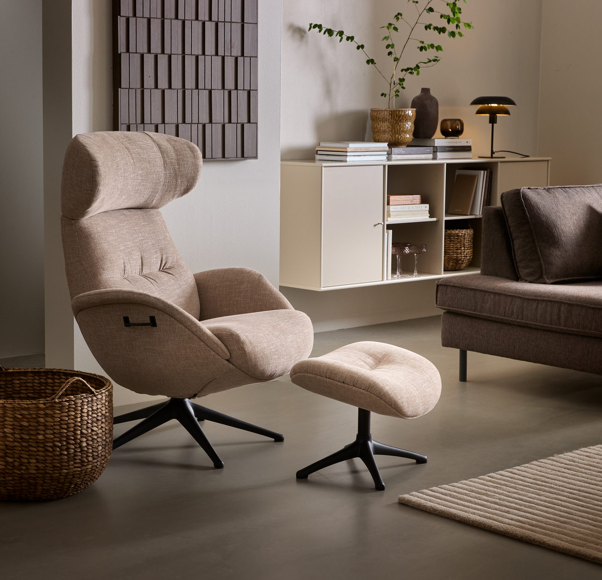 Beigefärgad loungefåtölj med matchande fotpall i ett vardagsrum med väggfast hylla, böcker, bordslampa, växt, soffa och korg i mjuka neutrala toner.