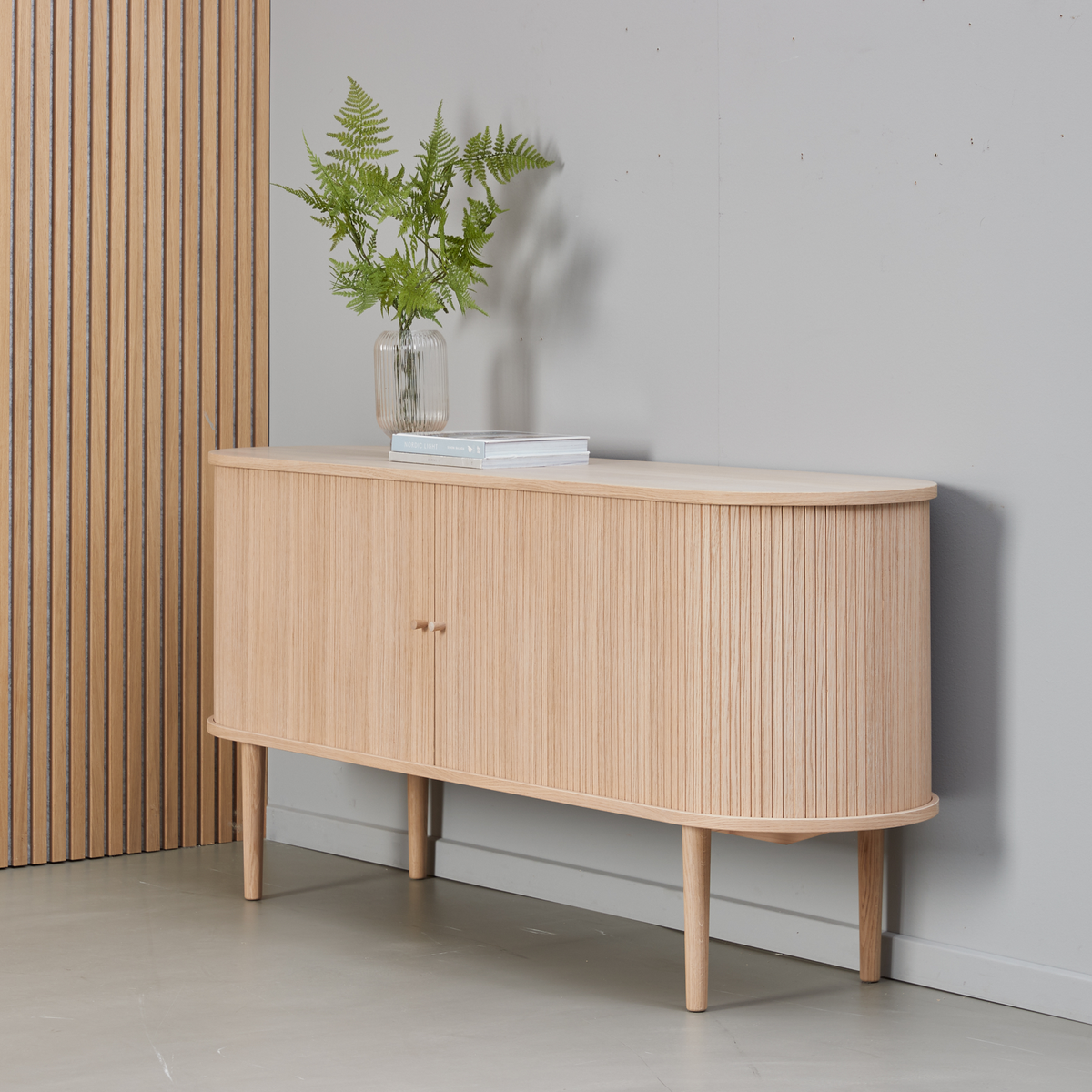 Sideboard i ljus ek med räfflade luckor på avsmalnande ben, dekorerat med glasvas med gröna ormbunkar och en bok mot en grå vägg.