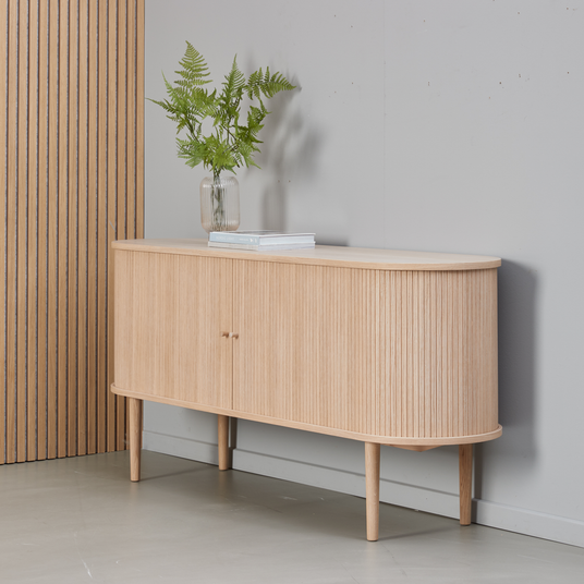 Sideboard i ljus ek med räfflade luckor på avsmalnande ben, dekorerat med glasvas med gröna ormbunkar och en bok mot en grå vägg.