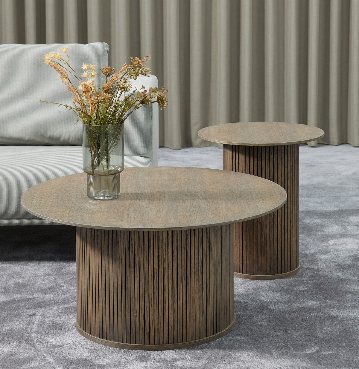Två runda soffbord i mörkt trä med räfflade cylinderunderreden; en glasvas med torkade blommor på det främre bordet, grå soffa, gardiner och grå matta.