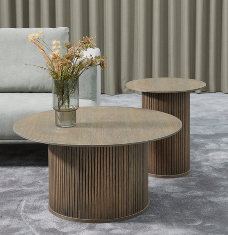 Två runda soffbord i mörkt trä med räfflade cylinderunderreden; en glasvas med torkade blommor på det främre bordet, grå soffa, gardiner och grå matta.