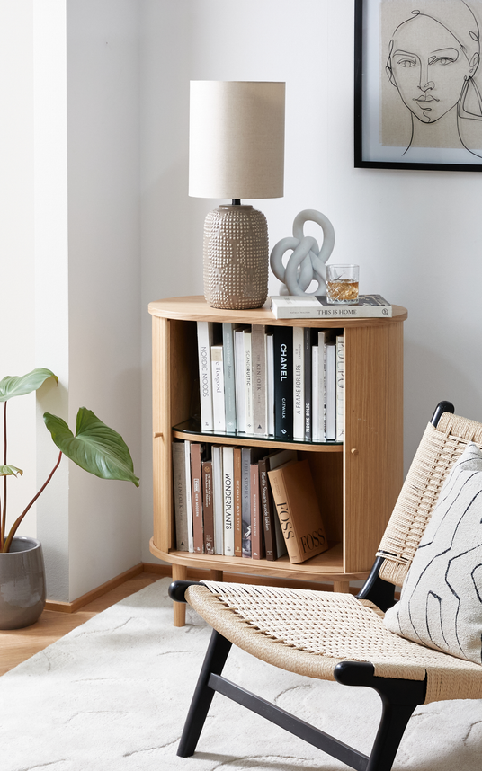Ljus skandinavisk läshörna med hörnhylla i trä fylld med designböcker; ovanpå står en beige bordslampa, en skulptural knut och ett glas med dryck på boken ”This Is Home”. En flätad loungestol med kudde, en pottväxt och en ljus matta kompletterar scenen; synliga boktitlar inkluderar ”CHANEL” och ”Nordic Moods”.