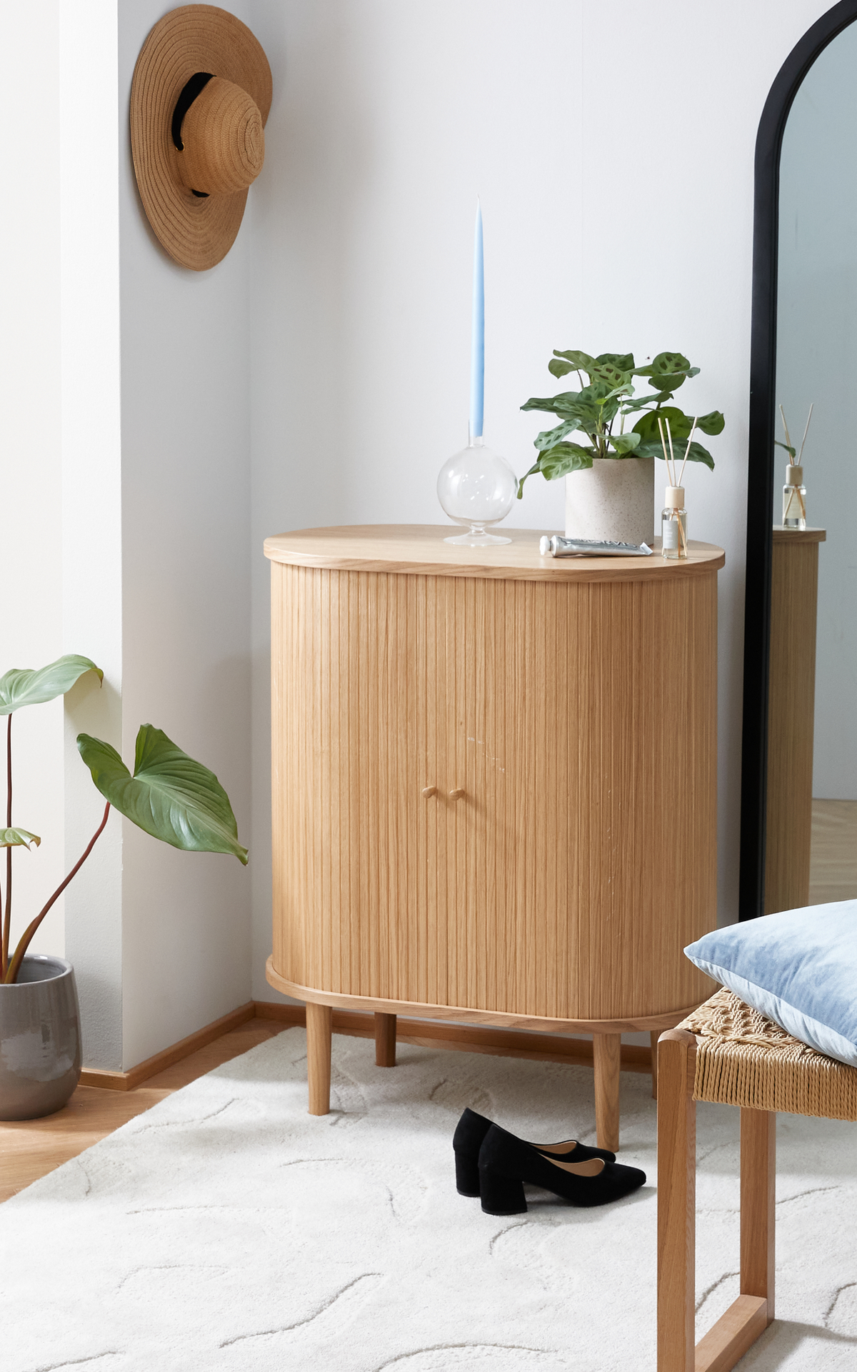 Ljus hall med rundat skåp i ljust trä, svartinramad spegel, krukväxt, ljusblåt ljus i glashållare och svarta klackar på vit matta.