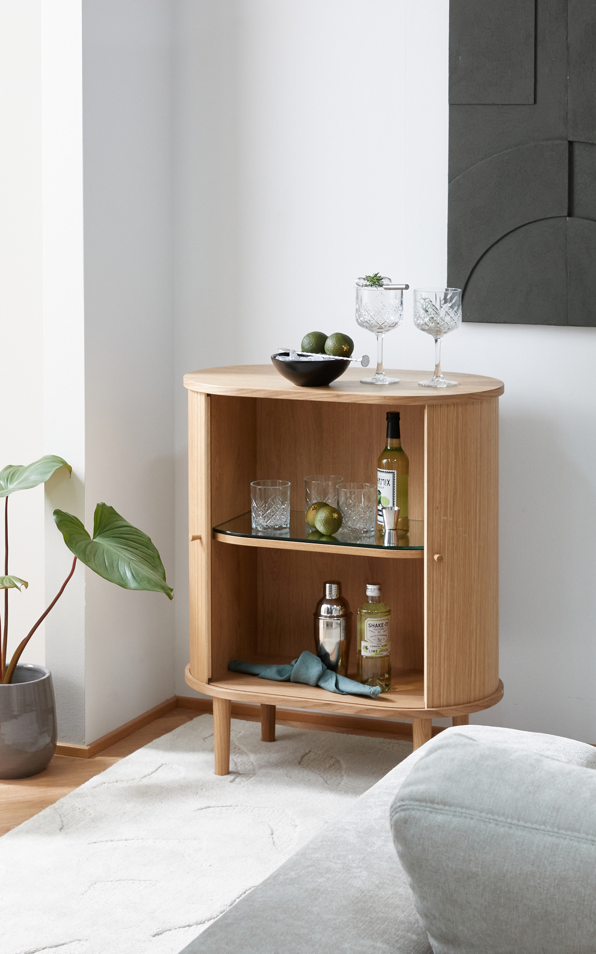 Litet rundat barskåp i ek med öppna hyllor, glas, shaker, flaskor och lime; två kristallglas och en skål med lime ovanpå, bredvid en krukväxt i ett ljust vardagsrum.
