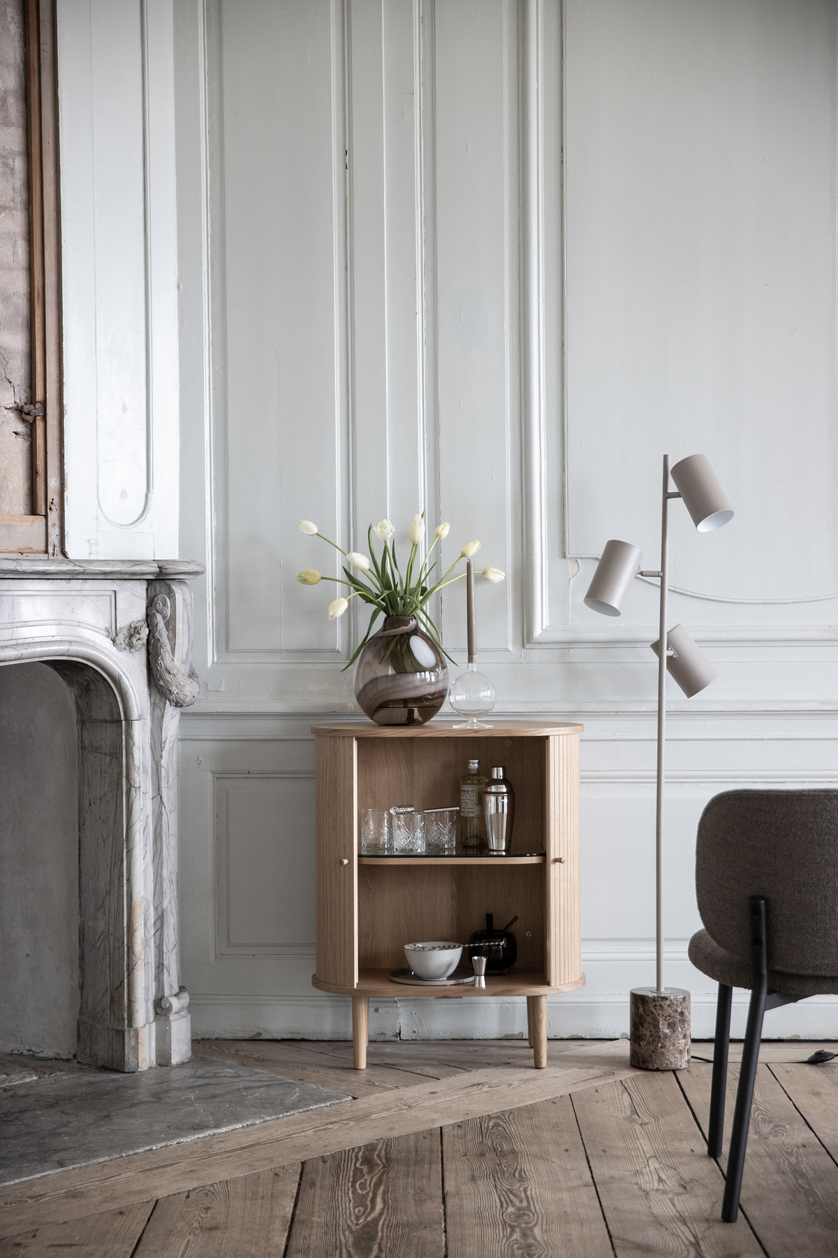 Ljus skandinavisk interiör med litet eksskåp, vas med vita tulpaner och en minimalistisk golvlampa vid en marmorspis framför en panelerad vägg.