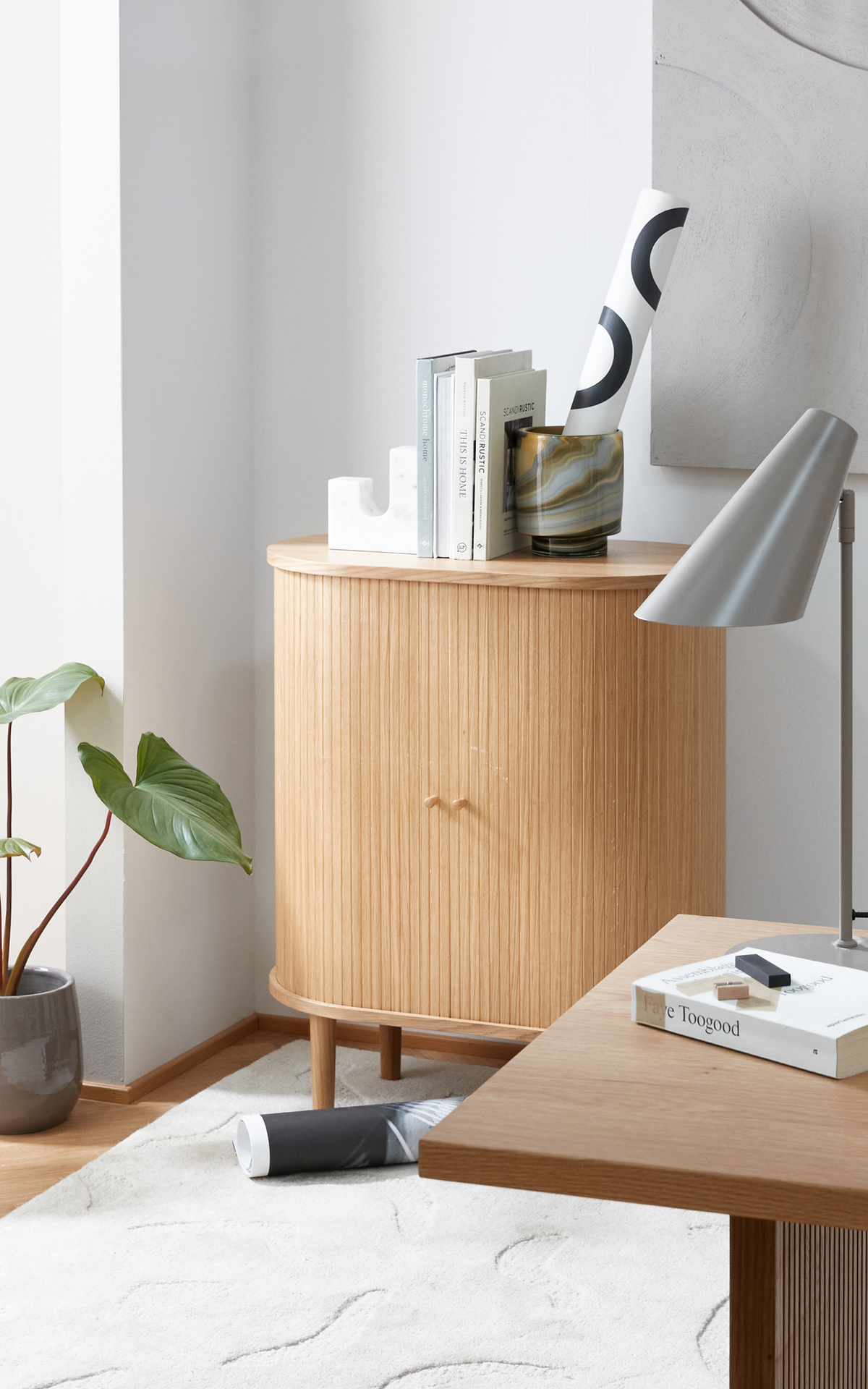 Ljus skandinavisk hörna med ljust träskåp i ribbad trä, skrivbord med grå bordslampa och en bok med titeln Faye Toogood; ovanpå skåpet står böcker, bokstöd och en mönstrad vas med rulle, samt en grön krukväxt och ljus matta.