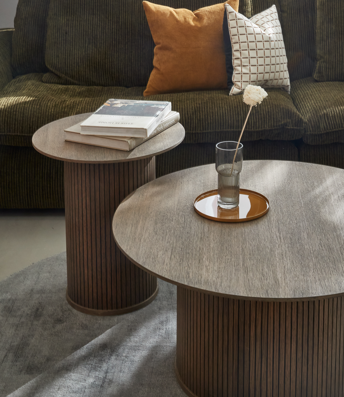 Två runda soffbord i mörk ek med räfflade sarger; på det ena ligger böcker, bland annat ”Nordic Interiors”, på det andra står en glasvas med en vit blomma på en brun bricka framför en olivgrön soffa med rostfärgad och vitrutigt kudde.