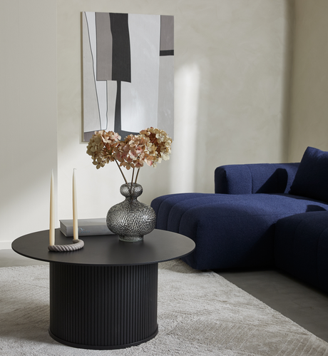 Minimalistiskt vardagsrum med svart runt soffbord med räfflad fot, glasvas med torkade hortensior, två cremefärgade ljus och en Dior-bok; mörkblå modulsoffa och abstrakt tavla på väggen.