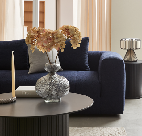 Mörkblå soffa med kuddar bakom ett runt soffbord med glasvas med torkade blommor, smal ljusljus och boken Dior; i bakgrunden skira gardiner och en metallbordslampa.