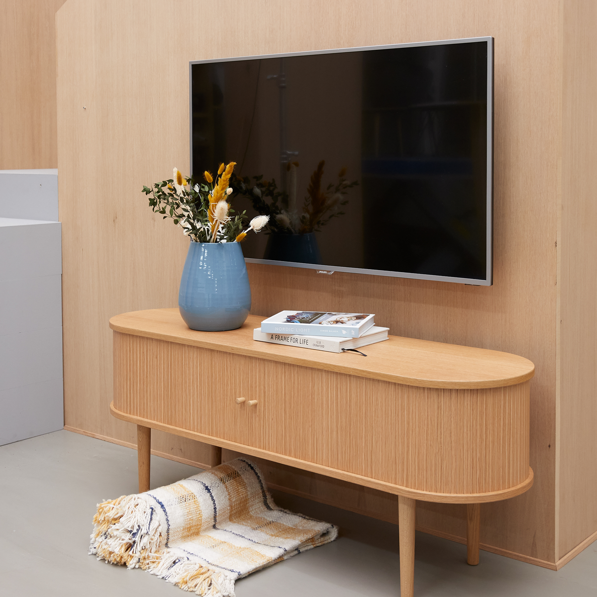Ljus trä-TV-bänk med blå vas med torkade blommor och böcker, inklusive ”A Frame for Life”, väggmonterad TV ovanför och en fransig pläd under bänken.