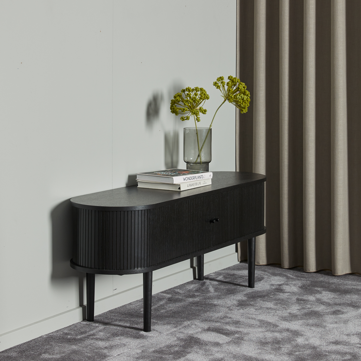 Svart ribbad sideboard mot ljusgrå vägg med vas med gröna stjälkar och två böcker, varav en med titeln “WONDERPLANTS 2”.
