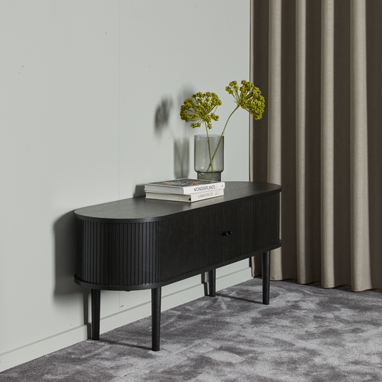 Svart ribbad sideboard mot ljusgrå vägg med vas med gröna stjälkar och två böcker, varav en med titeln “WONDERPLANTS 2”.