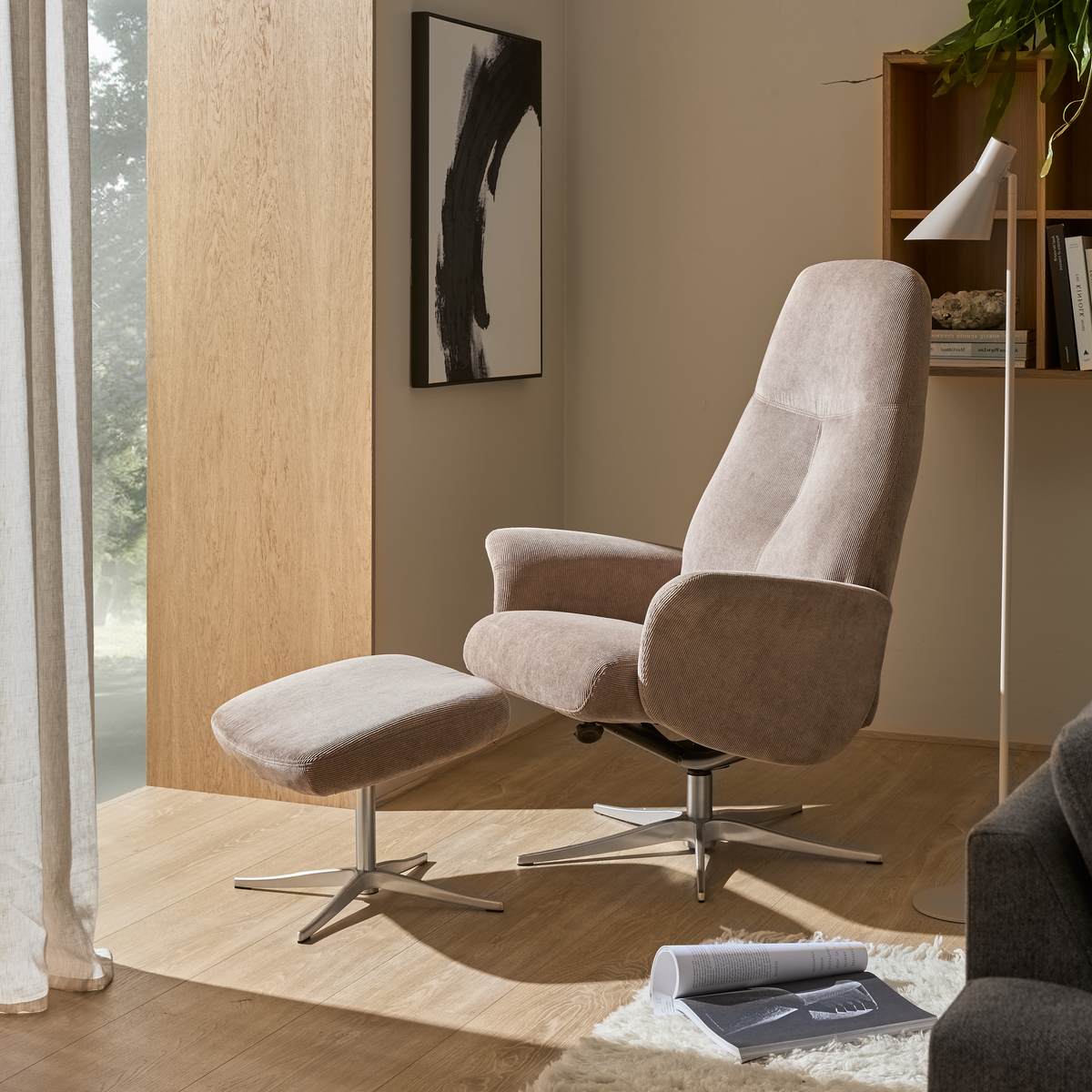 Beige recliner med matchande fotpall på metallfot i ett ljust vardagsrum vid fönster, med golvlampa, väggkonst och öppen hylla i dagsljus.