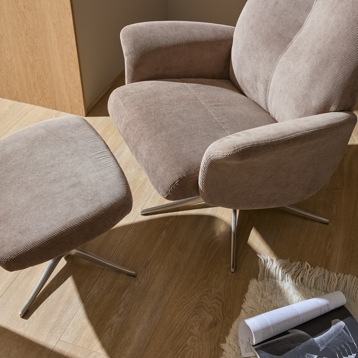 Beige fåtölj i manchester med matchande fotpall på trägolv, båda med metallben; solbelyst hörn med ljus ryamatta och uppslagen tidskrift på golvet.