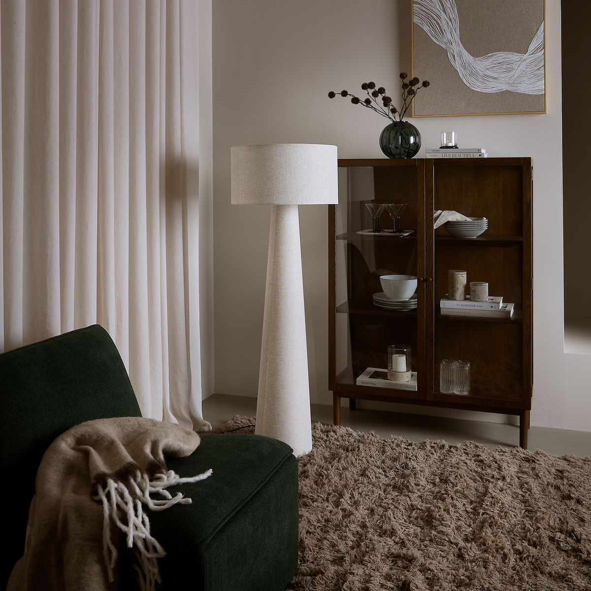 Mysigt vardagsrumshörn med mörkgrön fåtölj med beige pläd, hög vit golvlampa, ryamatta och vitrinskåp i mörkt trä med dukat porslin, böcker, vas och ljus.
