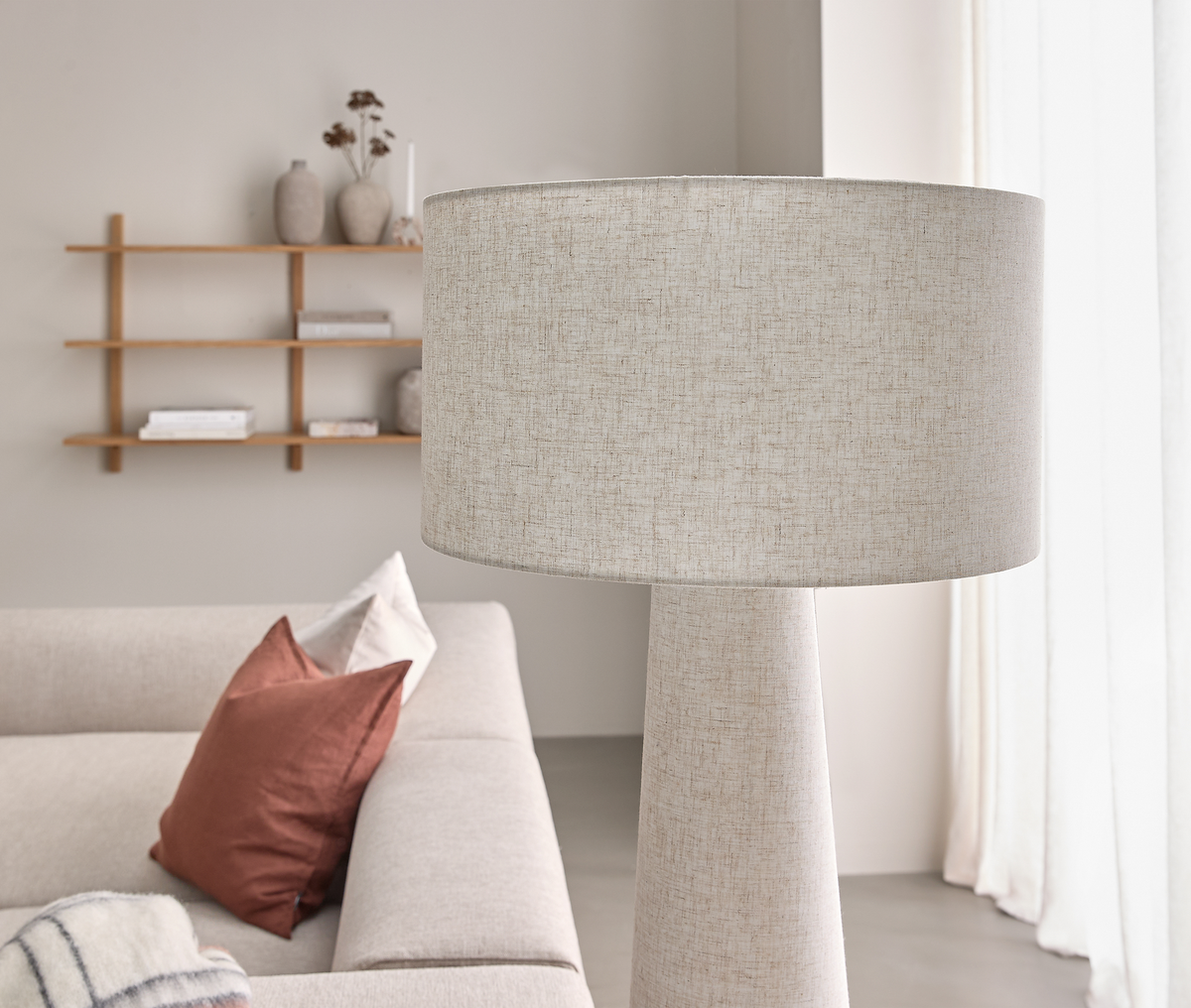 Beige golvlampa i tyg bredvid en ljus soffa med terrakottakuddar; hylla och ljusa gardiner i en skandinavisk vardagsrumsmiljö.