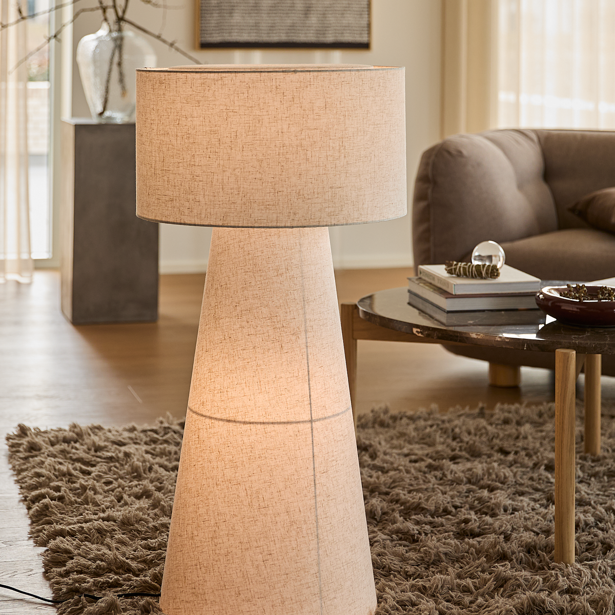 Stor golvlampa i beige tyg i ett vardagsrum med ryamatta, soffbord med böcker och en soffa.