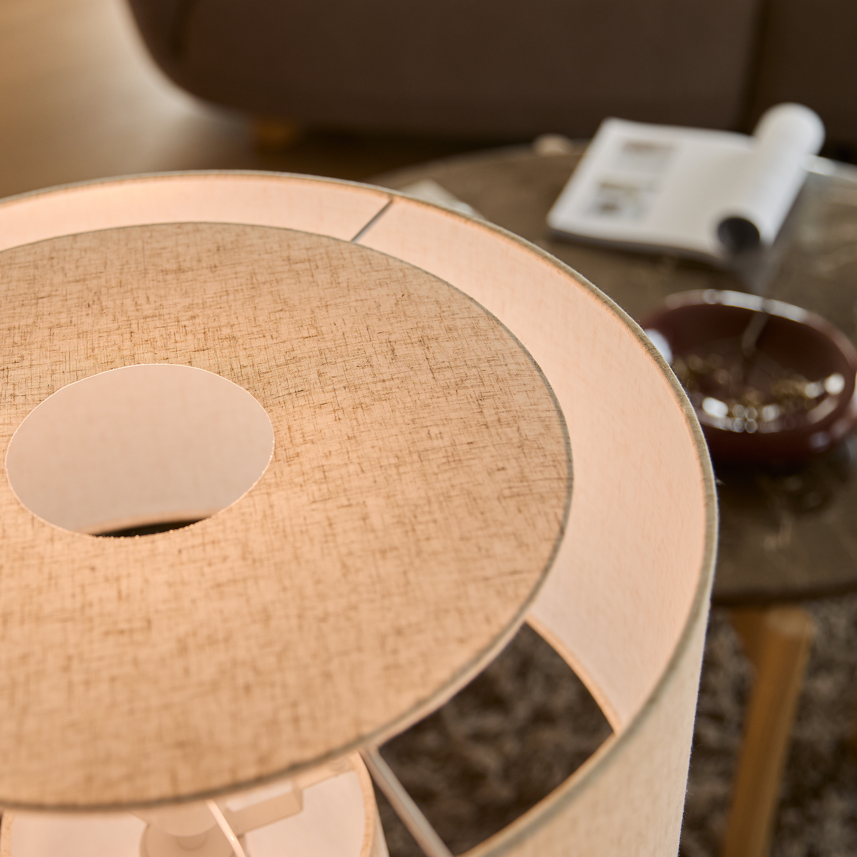 Närbild på beige textil lampskärm i vardagsrum, suddigt sofabord med opslagen tidning i bakgrunden.