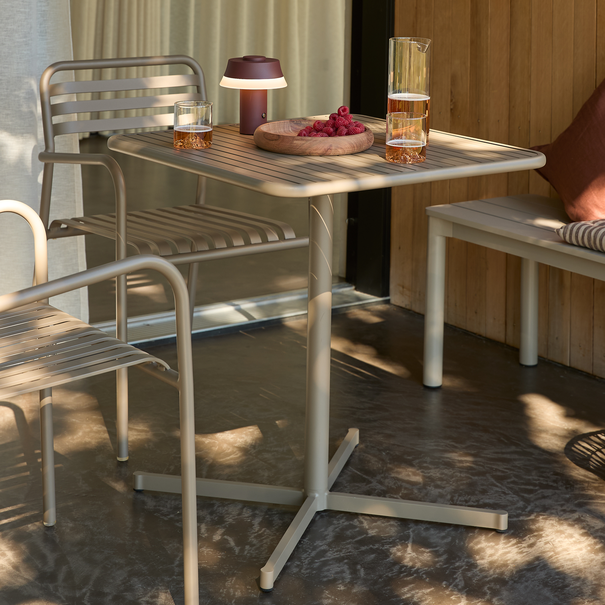 Beige utomhusmöbler på en terrass: ett fyrbent metallbord med två stolar, bänk med kudde, en liten portabel bordslampa, glas och karaff med dryck samt en trätallrik med hallon.