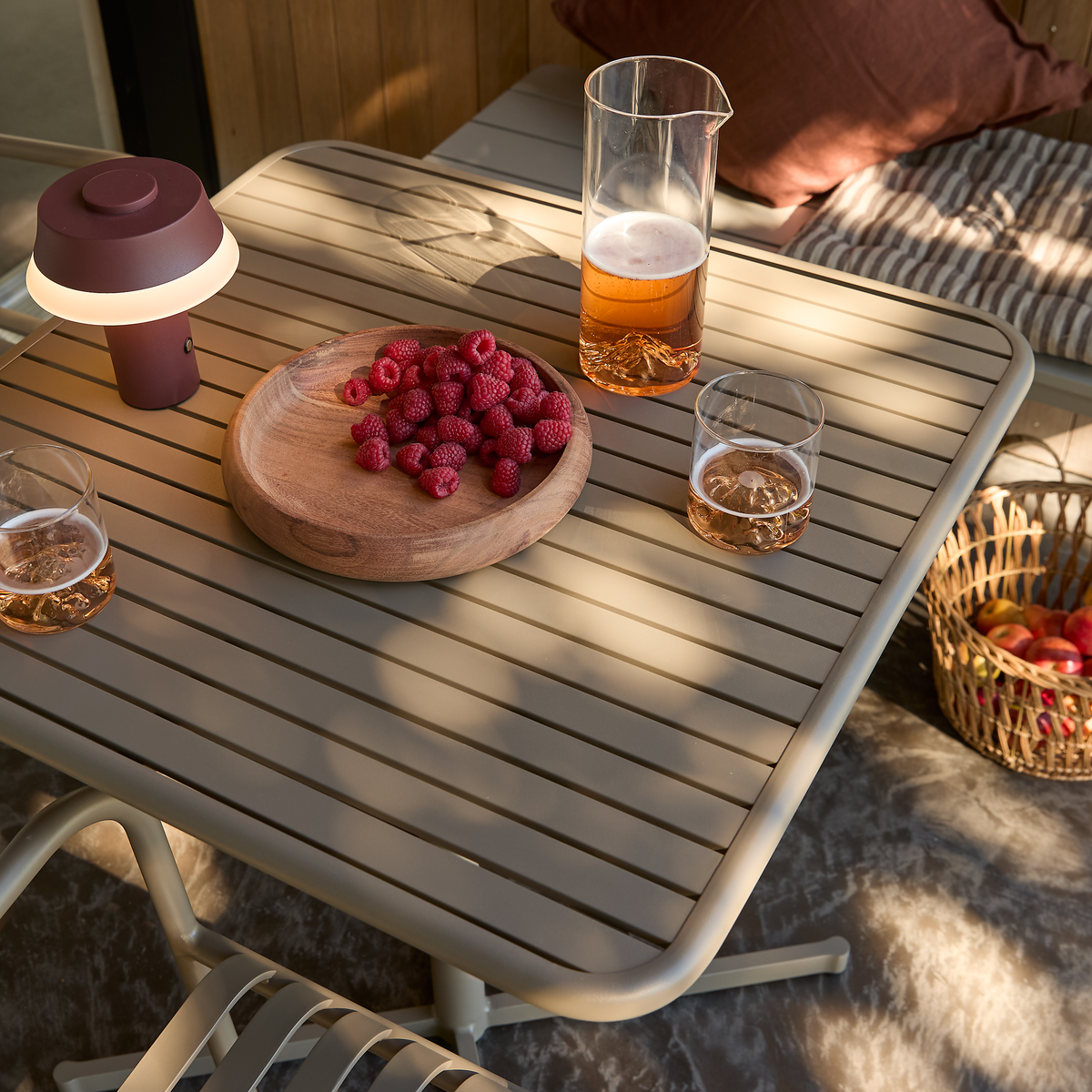 Terrassbord med träfat med hallon, kanna med roséfärgad dryck, två glas, portabel bordslampa och korg med äpplen i varmt solljus.