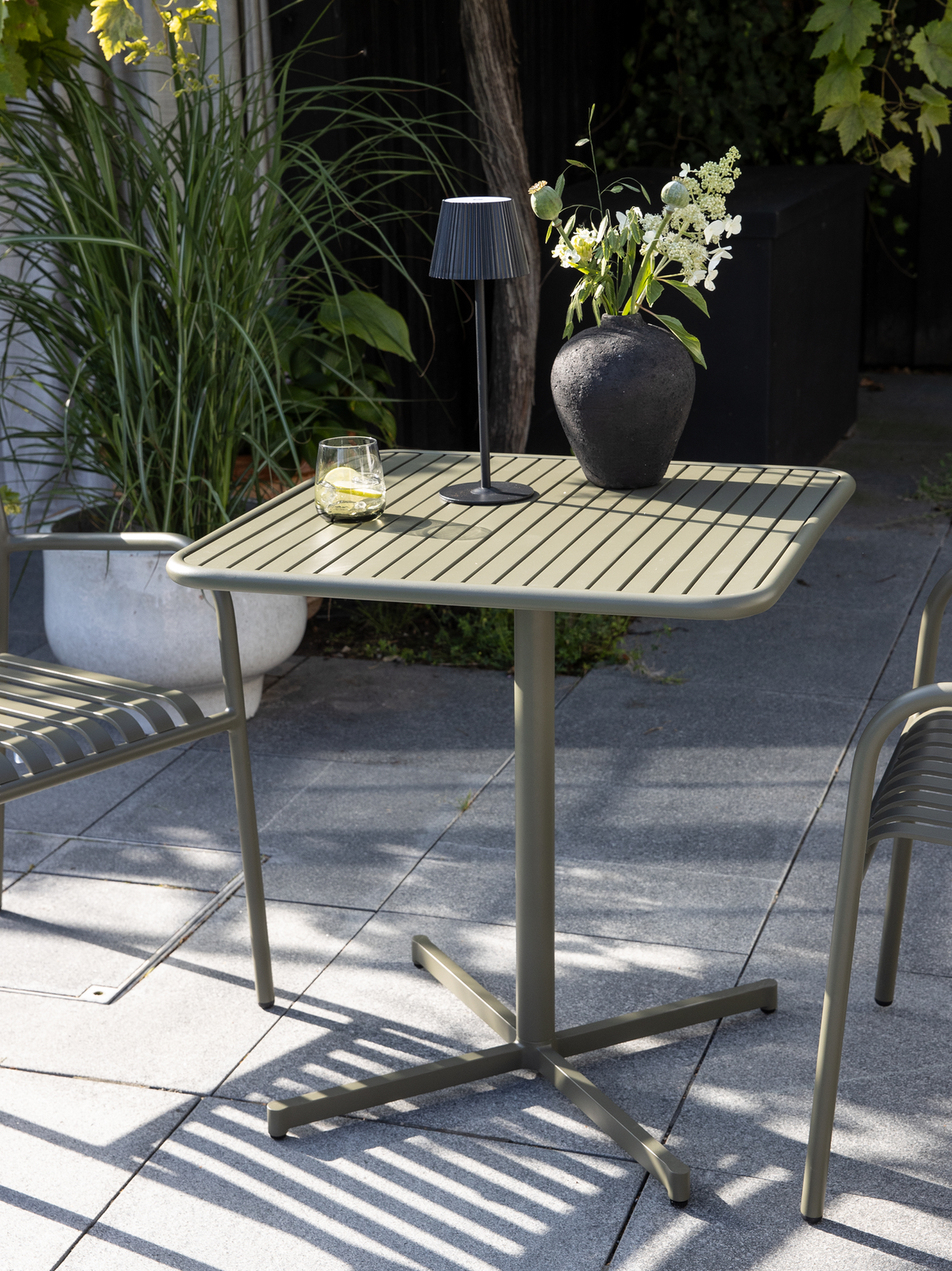 Olivgrönt utebord med ribbad skiva, liten svart bordslampa, vas med vita blommor och ett glas med citron på en grå terrass med matchande stolar och växter.