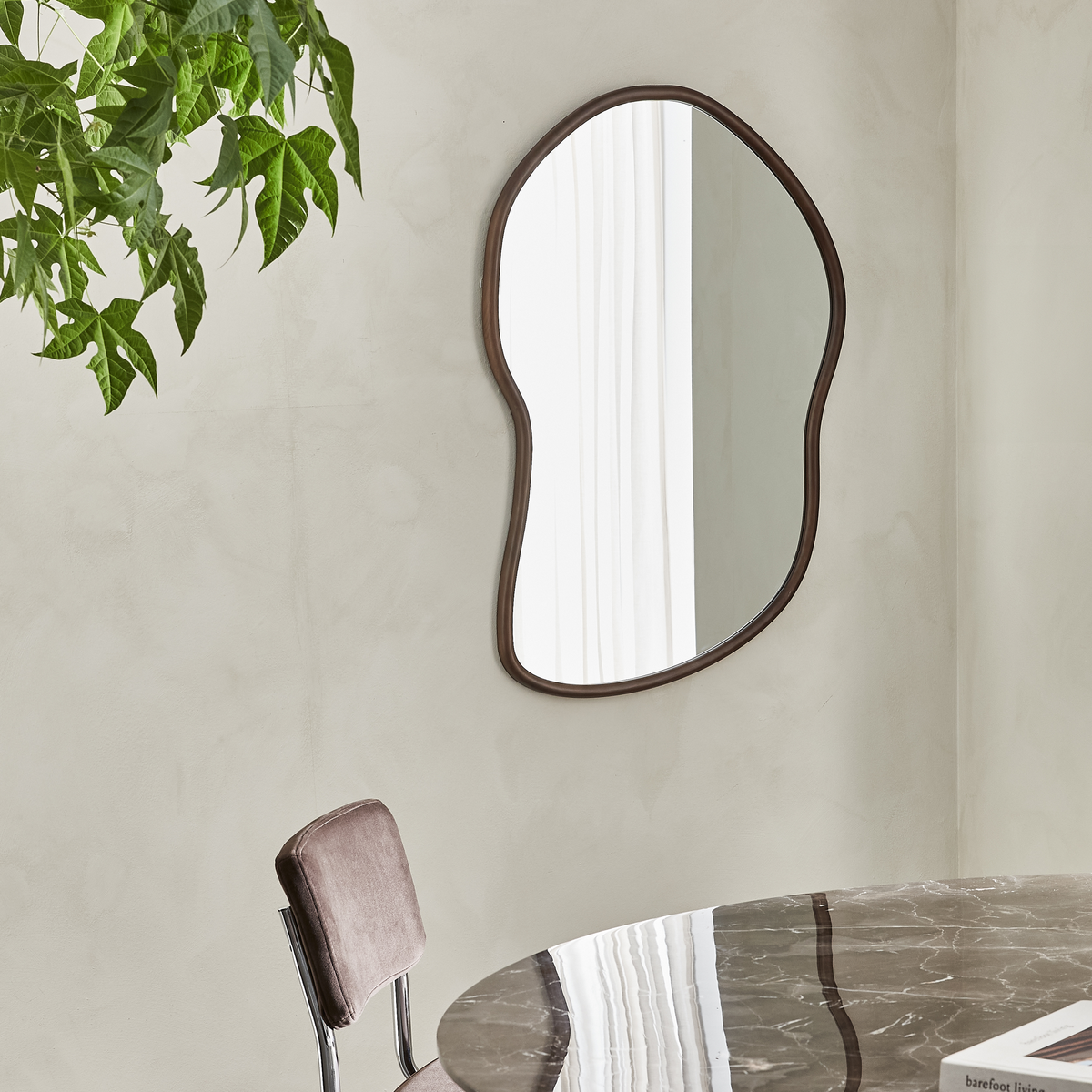 Organiskt formad spegel med mörk ram på beige vägg, vit gardin reflekteras; brun stol och marmorbord med boken ”barefoot living” i ett ljust matrum.