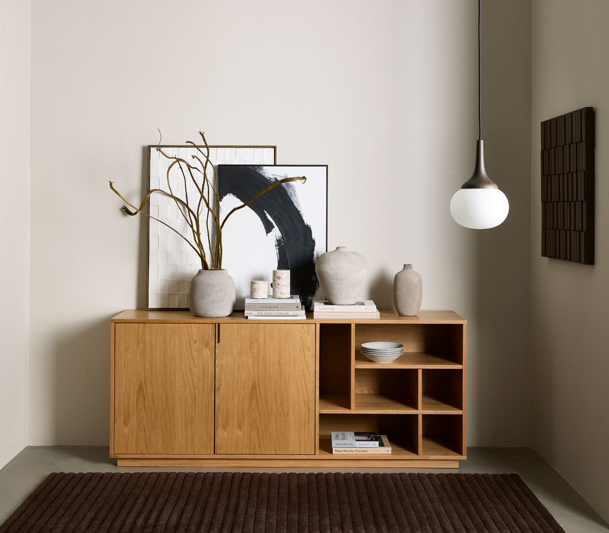 Minimalistisk hörna med ett sideboard i ek, neutrala vaser, två inramade konstverk, en hängande klotpendel och böcker – en bok har titeln “New Nordic Houses”.