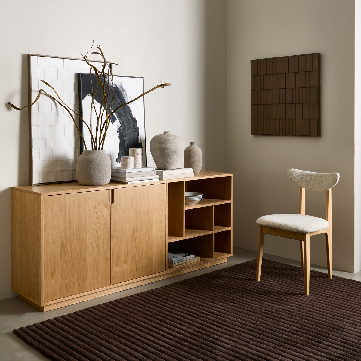 Ljus skandinavisk interiör med sideboard i ek, keramikvaser med torkade grenar och abstrakt konst, intill en trästol med ljus sits och en ribbad mörkbrun matta.