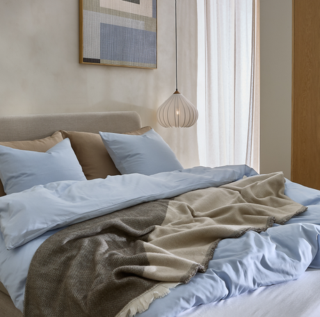 Ljust sovrum med bäddad säng i ljusblått påslakanset, beige kuddar och en beige‑grå filt, hängande vit pendellampa, gardiner och abstrakt väggkonst i mjuka neutrala toner.