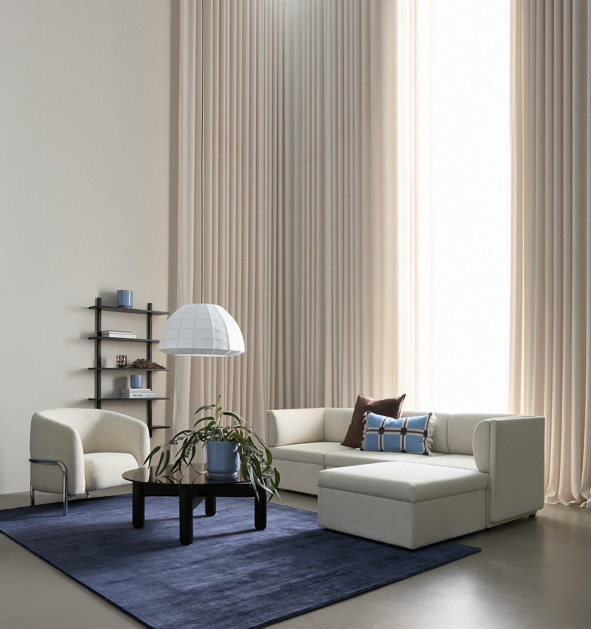 Ljust vardagsrum med beige modulsoffa, blå matta, svart soffbord med grön växt, pendellampa och höga beige gardiner.